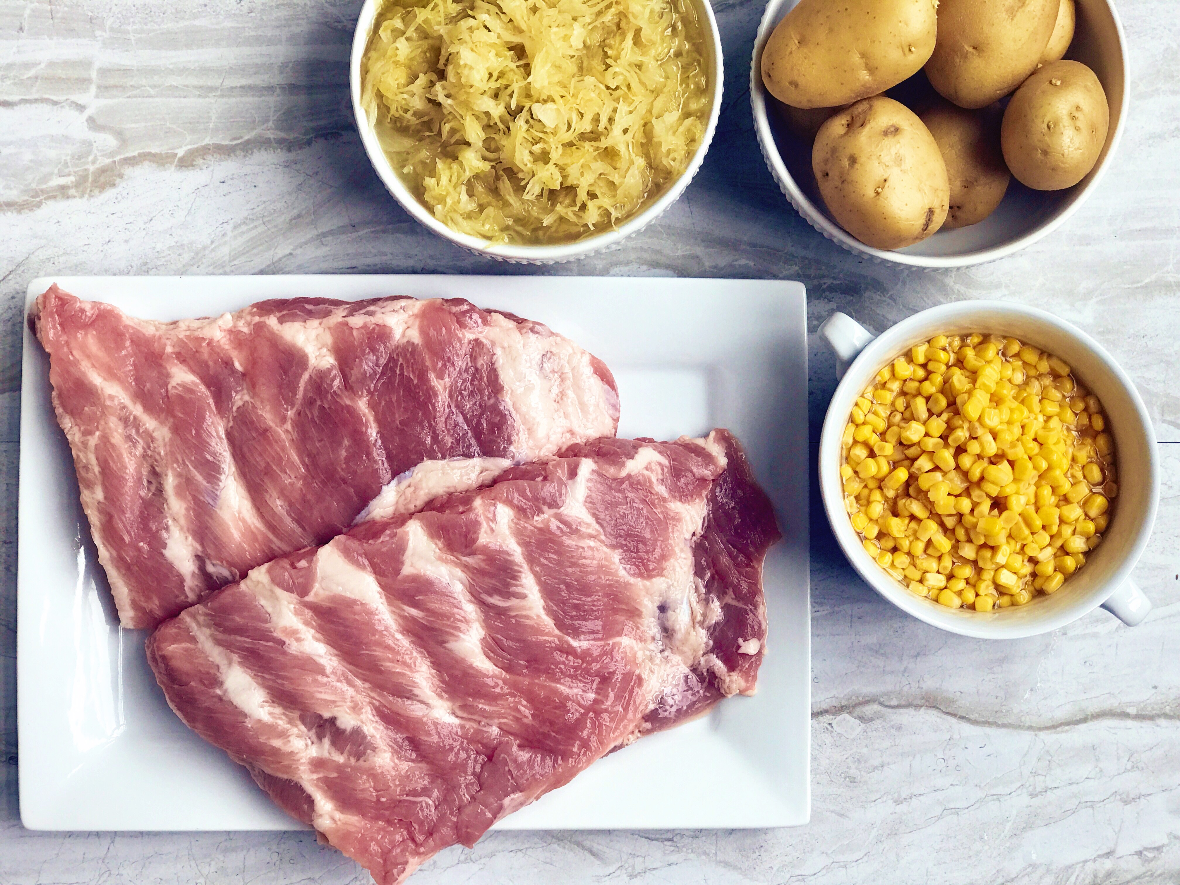 An image of some of the ingredients needed to make German Sauerkraut and Spareribs: pork spare ribs, corn, potatoes, and sauerkraut. 